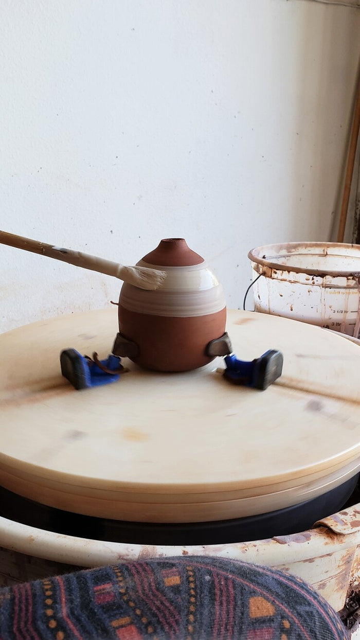 Brushing a white band onto a red clay pot held by blue Giffin Grip sliders on a spinning wheel