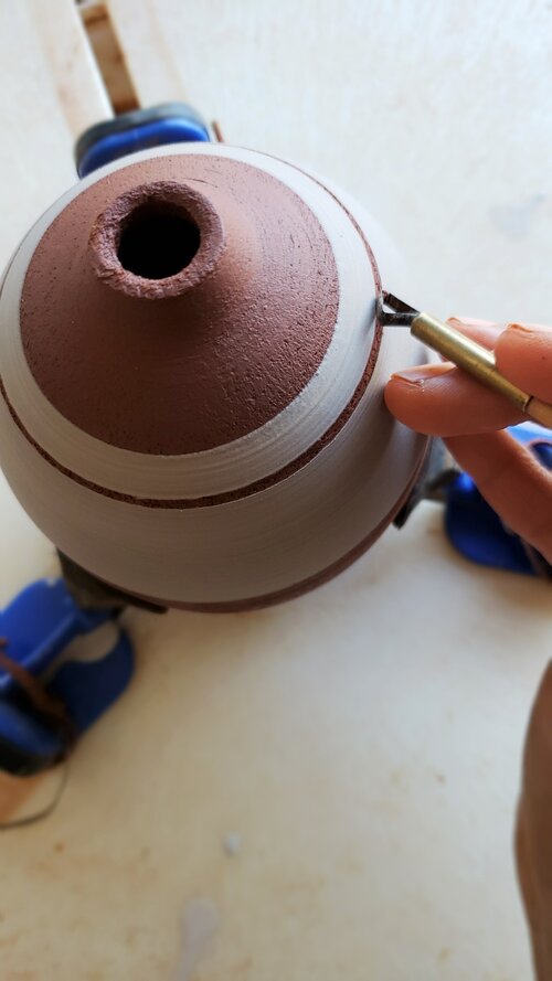 Carving bands through a brown coating on a bottle-necked greenware pot held by blue Giffin Grip sliders